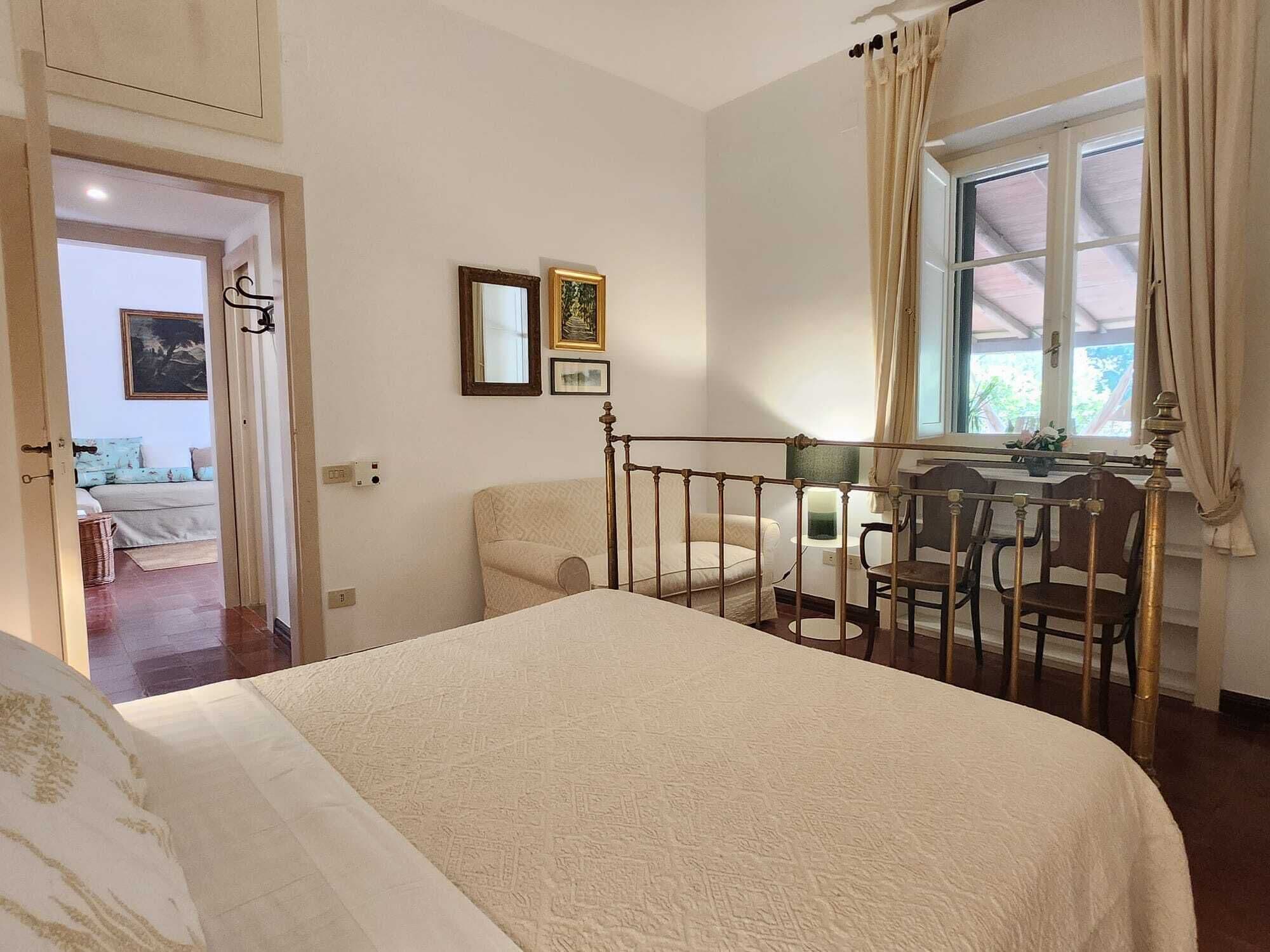 Quarto com cama, escrivaninha e cadeiras, janela com cortinas e porta de acesso a outro cômodo.
