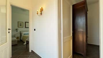 Ein Flur mit einer geschlossenen Holztür, einer weißen Wand mit Wandleuchte und einem Bett im angrenzenden Zimmer.