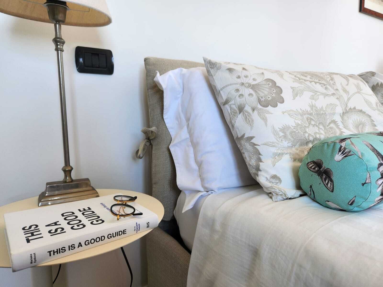 A neatly made bed with a floral pillow and a teal pillow with a butterfly pattern. A book titled "THIS IS A GOOD GUIDE" is placed on a bedside table next to a lamp.