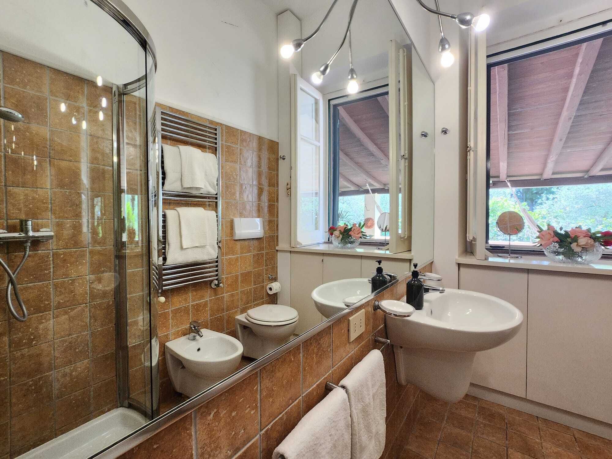 A modern bathroom with a glass shower enclosure, two sinks, a bidet, and a large mirror.