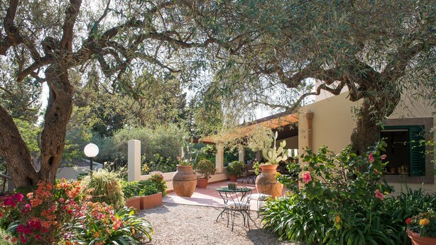 An outdoor patio area with a gravel path, potted plants, a table with chairs, and a building with a green door. - Don Gaspano  (Pace del Mela)