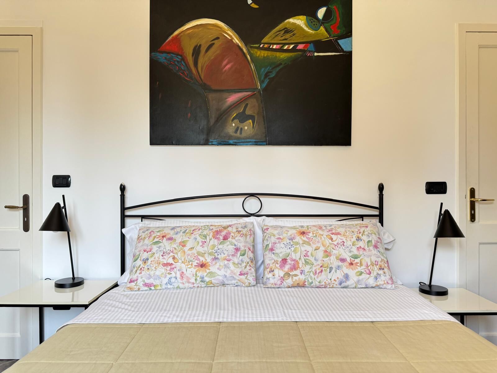 A bedroom with a bed, two nightstands, a painting above the bed, and a door on the left.