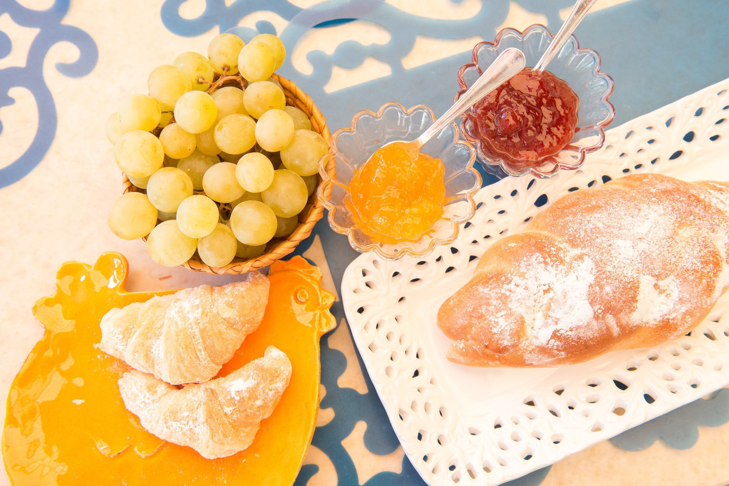 A table with a basket of yellow grapes, a bowl of orange jam, a plate with pastries, and a bread roll.