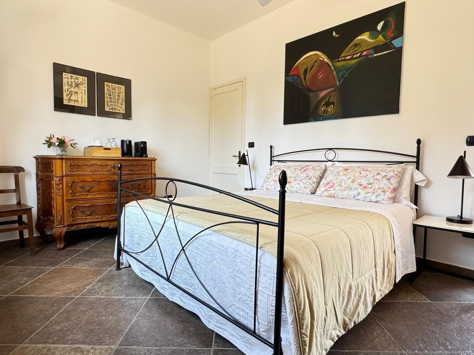 A bedroom with a metal bed frame, a wooden dresser, a nightstand, a lamp, and a painting on the wall.