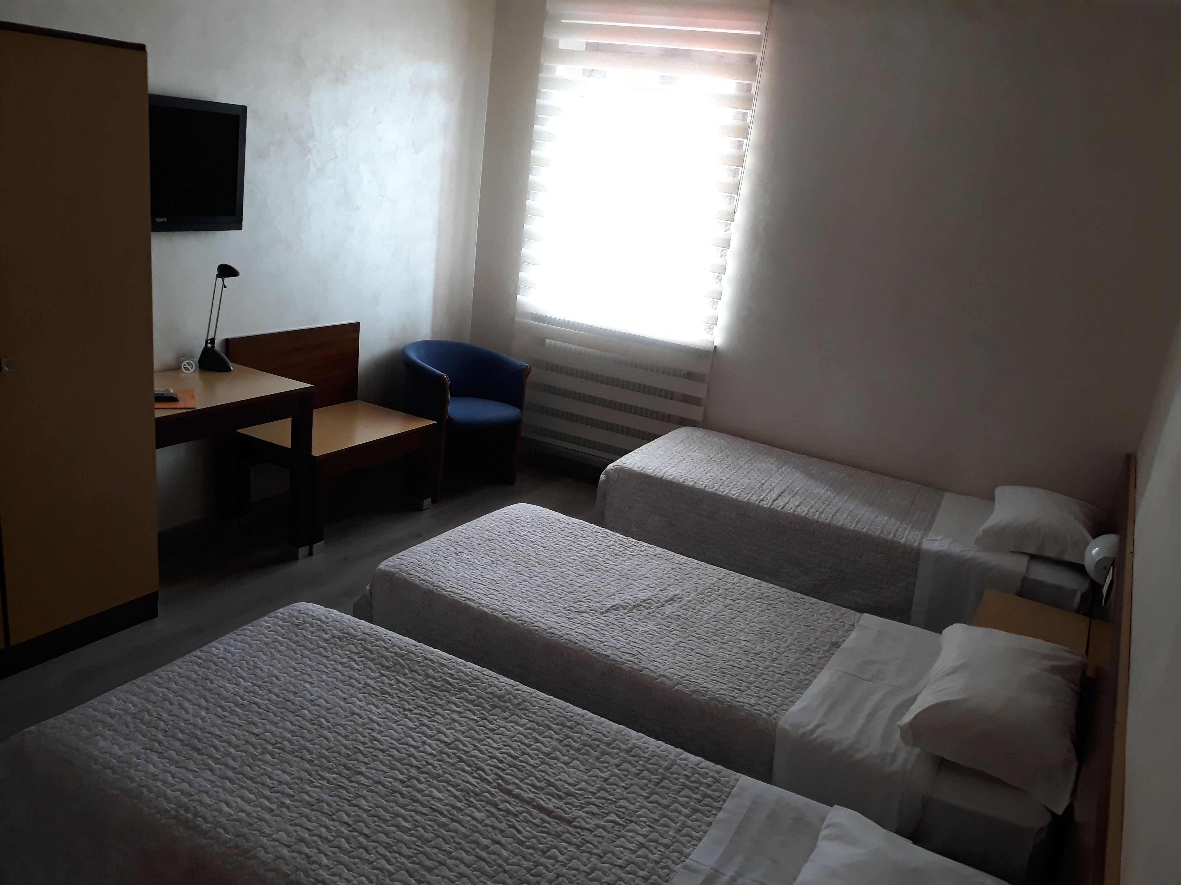 A hotel room with two single beds, a desk with a chair, a TV mounted on the wall, and a window with blinds.