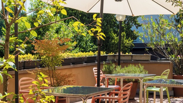Een terras buiten met tafels en stoelen, omringd door groen en een grote parasol voor schaduw.