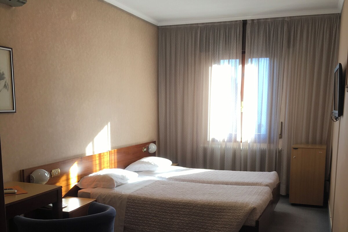 A hotel room with two beds, a desk, a chair, a small cabinet, and a window with curtains.