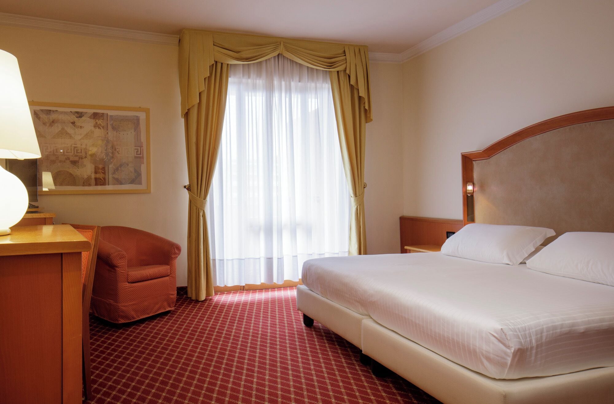 Une chambre d’hôtel avec un lit, une chaise, un bureau, une lampe et une fenêtre avec des rideaux.