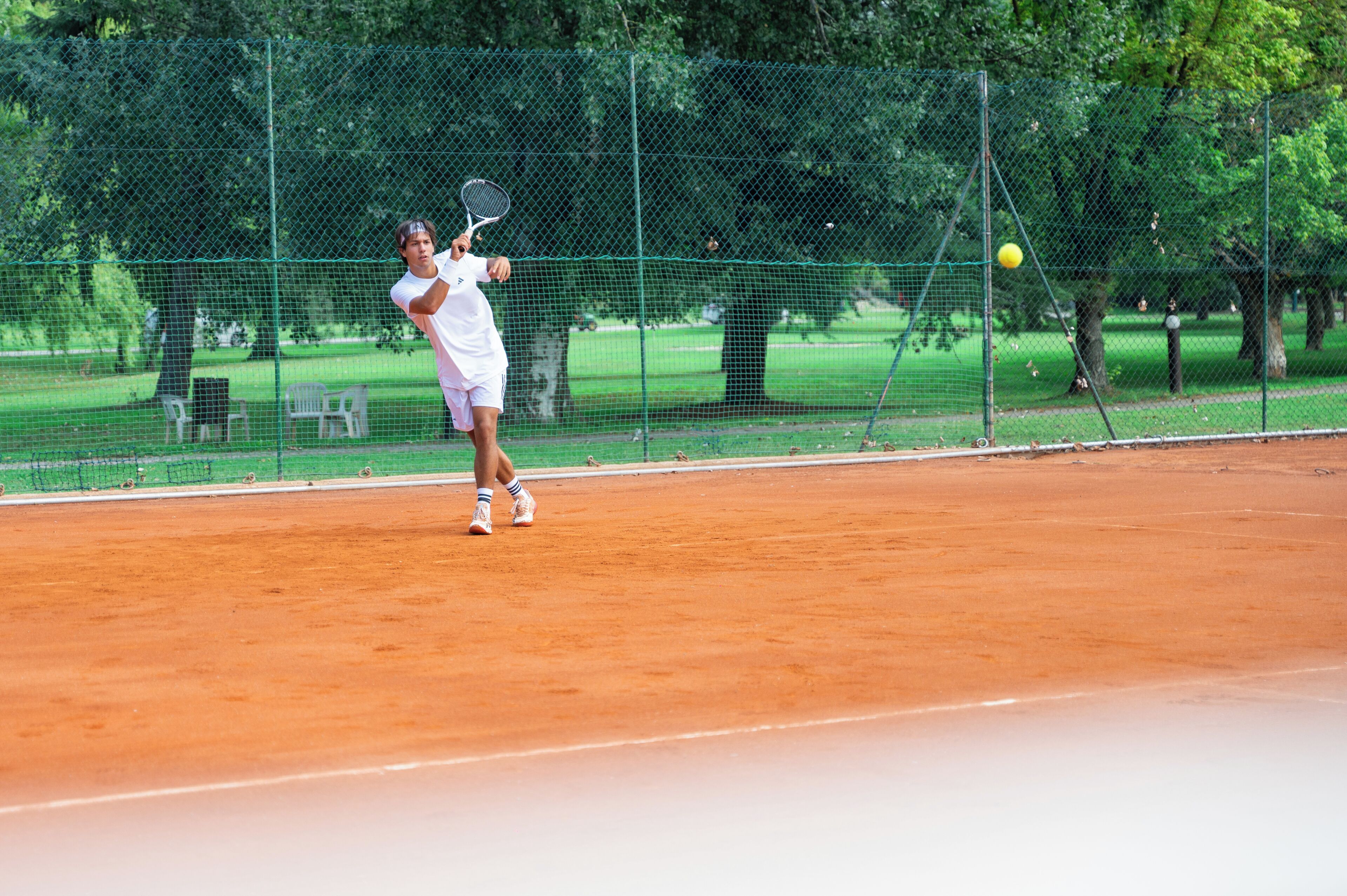 A tennis court with a player in action, surrounded by a green fence and trees.