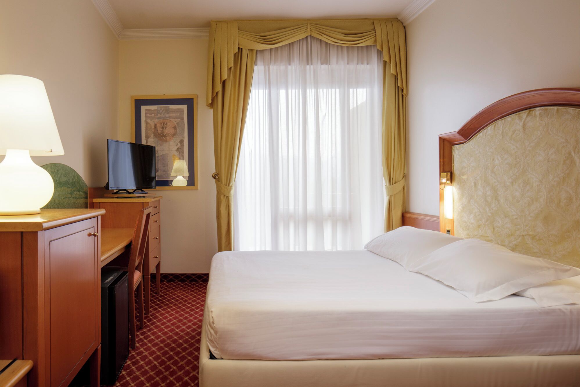Una camera d'albergo con un letto, una scrivania con una lampada, una televisione e un quadro appeso al muro.
