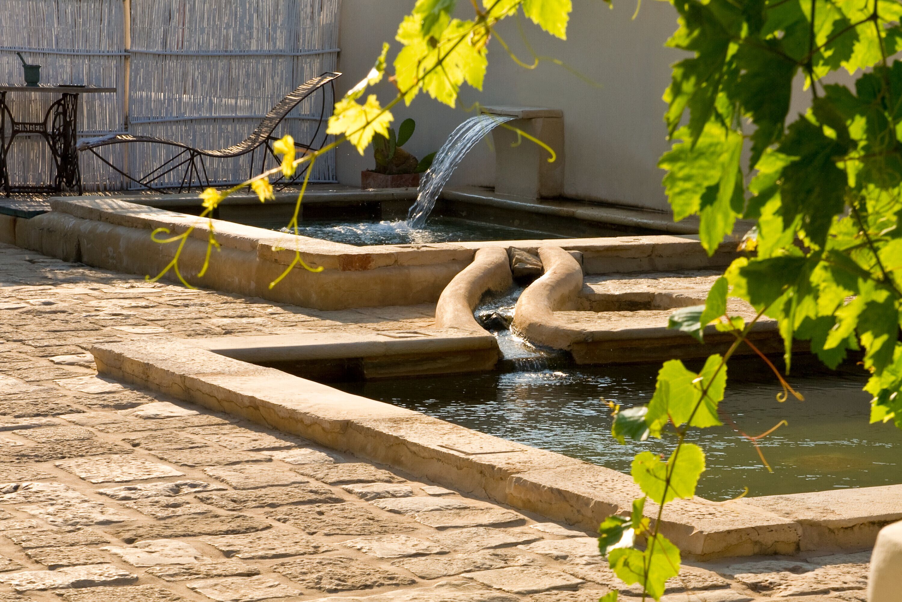 A stone fountain with water flowing into a rectangular basin, surrounded by a cobblestone courtyard.