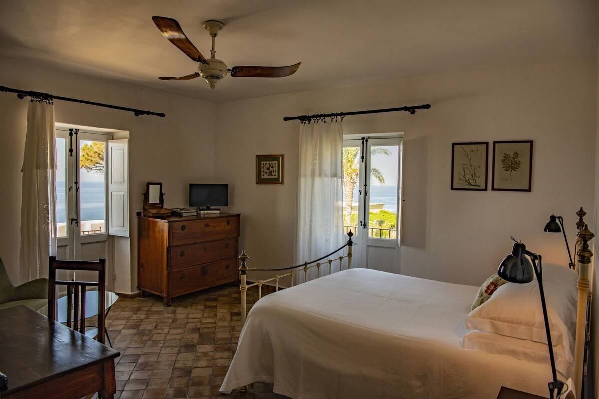 Un dormitorio con cama, escritorio, cómoda, ventilador de techo y ventana con vista al mar.