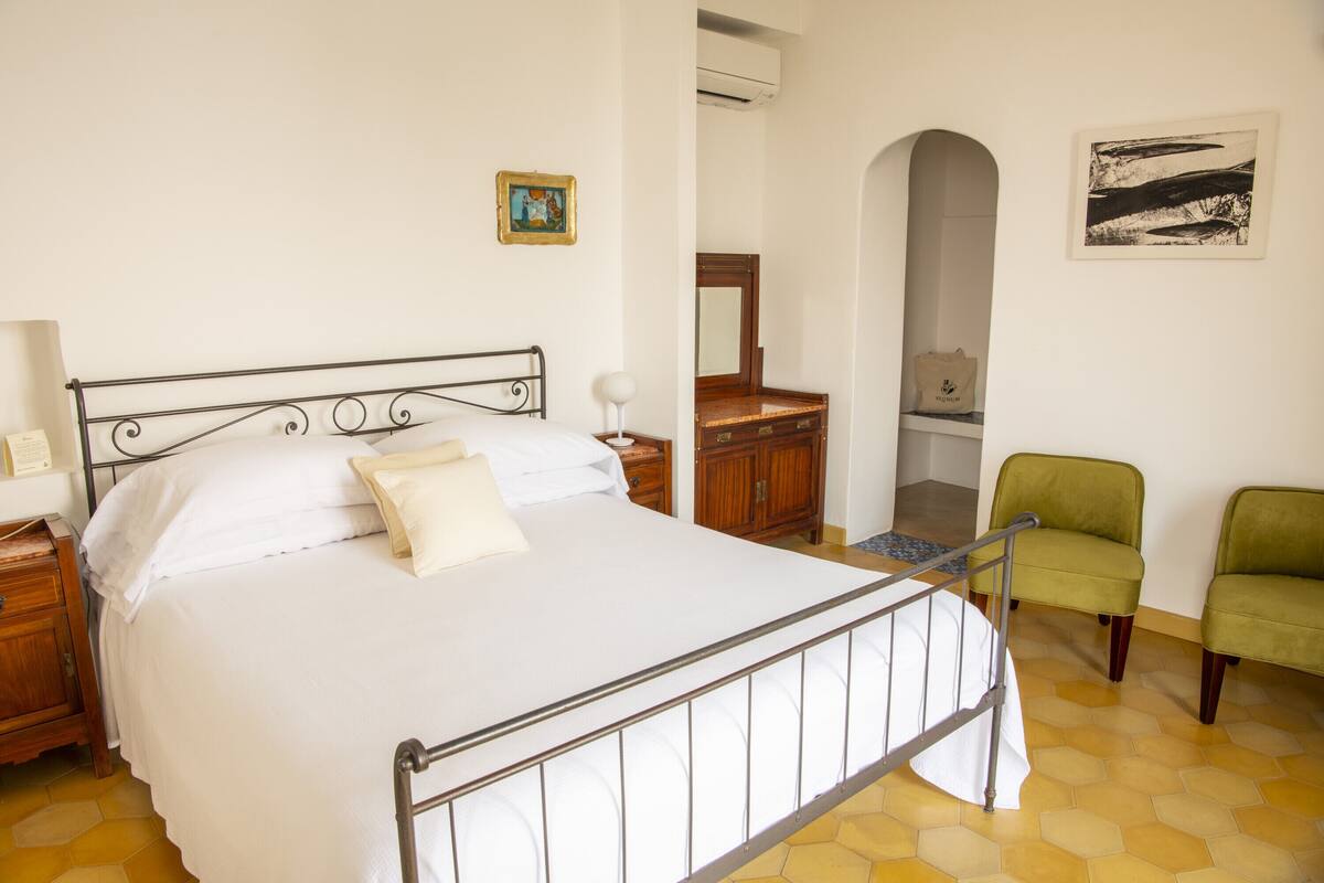 Un dormitorio con una cama de estructura metálica, un aparador de madera, un sillón verde y un espejo.
