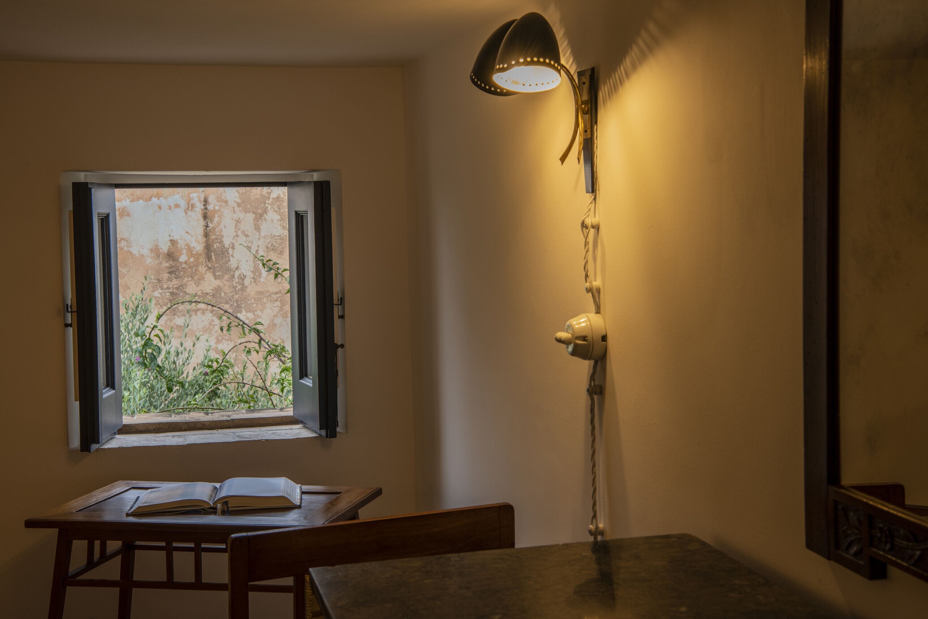 Una stanza con un tavolo, un libro aperto, una finestra con vista sul verde e una lampada fissata al muro.