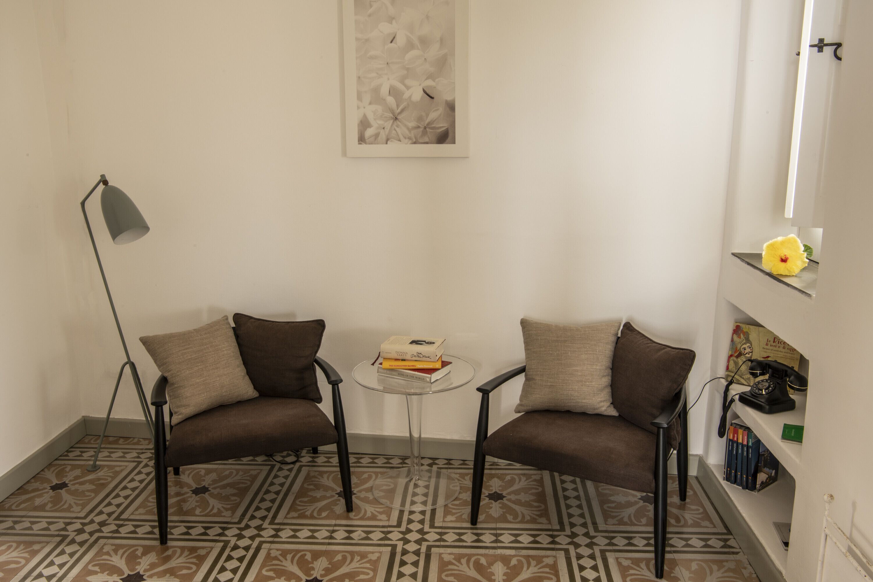 Ein Zimmer mit zwei Sesseln, einem kleinen runden Tisch, einer Stehlampe und einem Bücherregal mit Büchern und einer Vase.