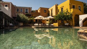 A clear pool with lounge chairs and umbrellas, surrounded by buildings with balconies and steps leading to the pool.
