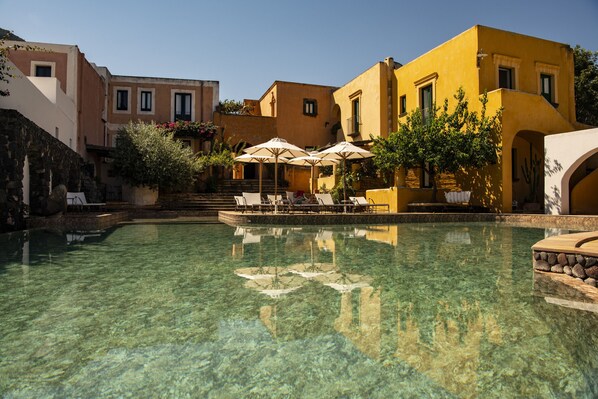A clear pool with lounge chairs and umbrellas, surrounded by buildings with balconies and steps leading to the pool.