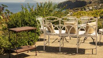A patio with a white metal table and chairs, surrounded by greenery and overlooking a coastal landscape.