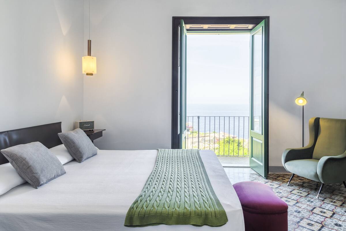 Una habitación de hotel con una cama, una mesita de noche, una silla y vistas al exterior.