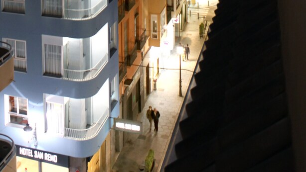 A hotel with multiple balconies, a visible entrance, and a street view with buildings and a mountain in the background. - Hotel San Remo (Alicante)