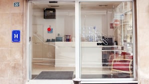 The entrance to Hotel San Remo with a clear glass door, a reception desk inside, and a blue sign indicating the presence of a handicap-accessible room. - Hotel San Remo (Alicante)