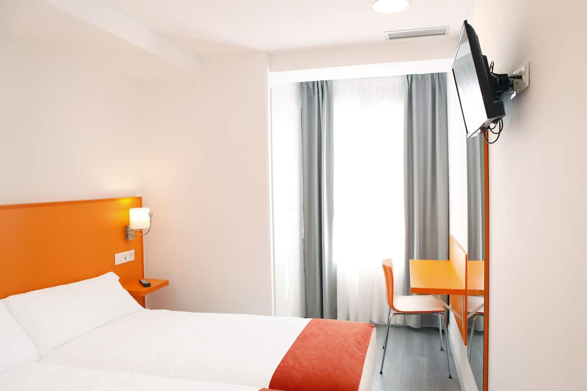 A hotel room with a bed, a desk, a chair, a TV mounted on the wall, and a window with curtains.