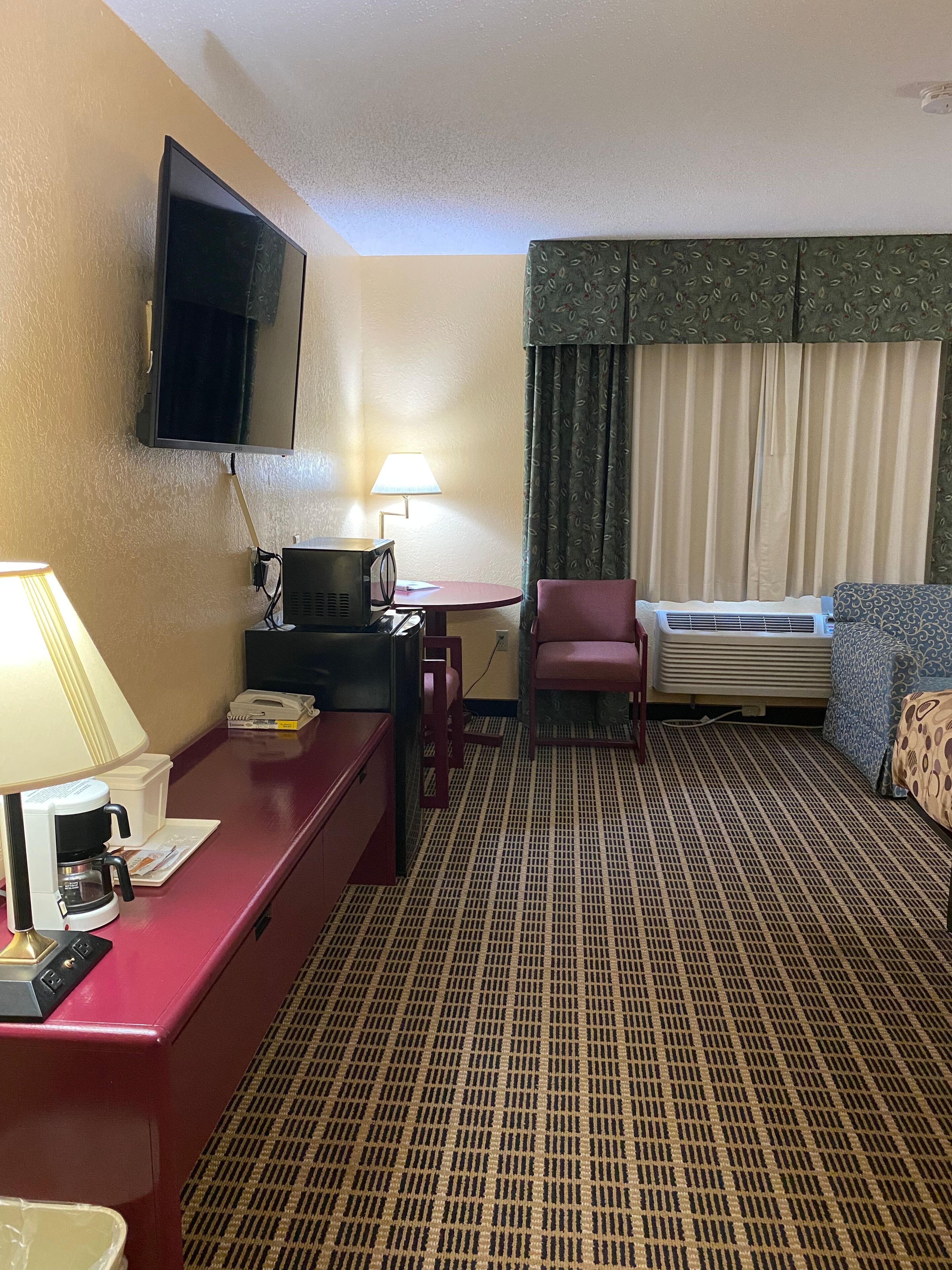 A hotel room with a desk, chair, television, air conditioning unit, and patterned carpet.
