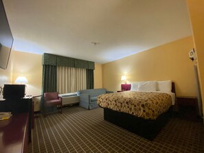 Une chambre d’hôtel comprenant un lit, un bureau avec un ordinateur, une chaise, un canapé, une fenêtre avec des rideaux et une table de chevet avec une lampe.