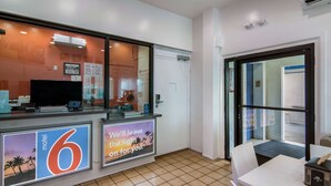 Zona de recepción de un hotel con un letrero digital que muestra el número 6, un televisor y una puerta de cristal que da al exterior.
