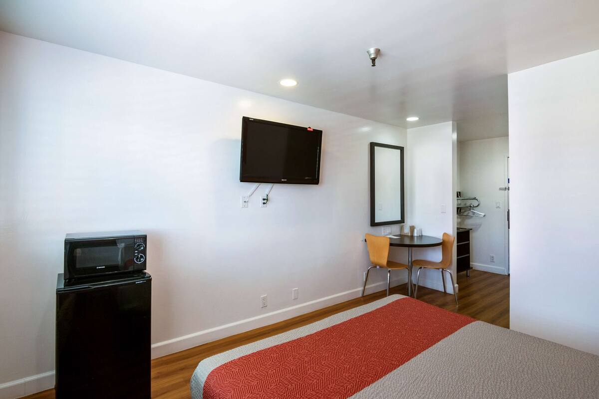 A compact hotel room with a bed, a microwave, a TV mounted on the wall, and a small dining area with a table and chairs.