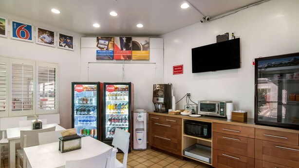 Un área de cocina limpia y bien iluminada, con refrigerador, microondas y cafetera. Hay mesas y sillas blancas, y un ventanal grande a la izquierda.