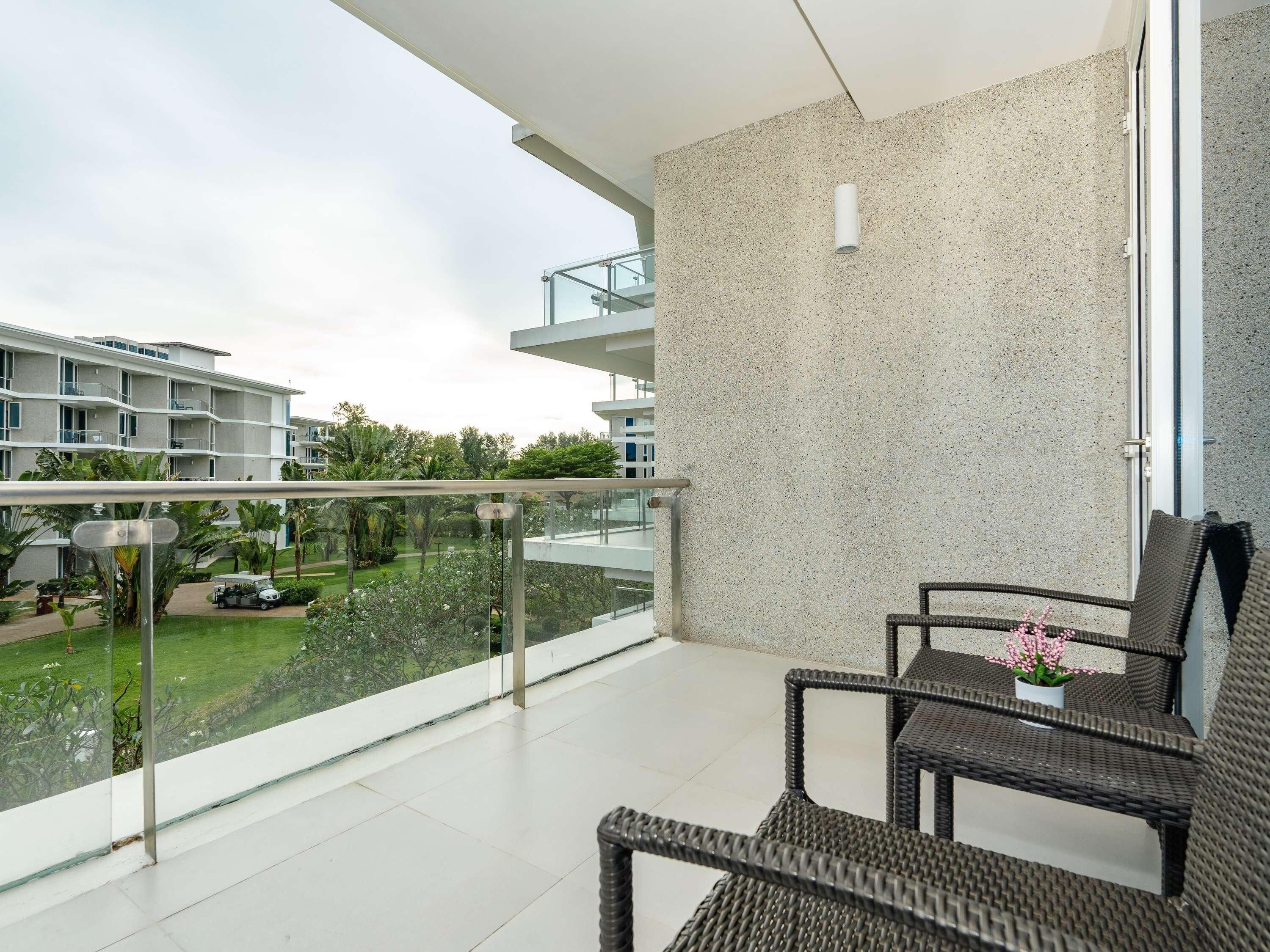 A balcony with wicker furniture, a glass railing, and a view of a residential building and greenery.
