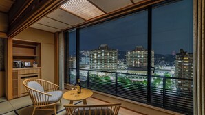 A balcony with tatami mats, a wooden chair, a small table, and a view of city buildings at night. - Hotel Wakamizu (Takarazuka)