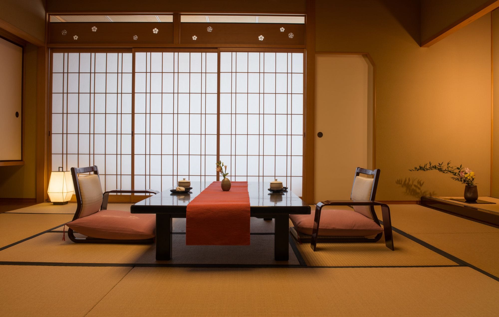 A traditional Japanese room with tatami flooring, a low table with a red runner, two chairs, a tea set, and a shoji screen door.