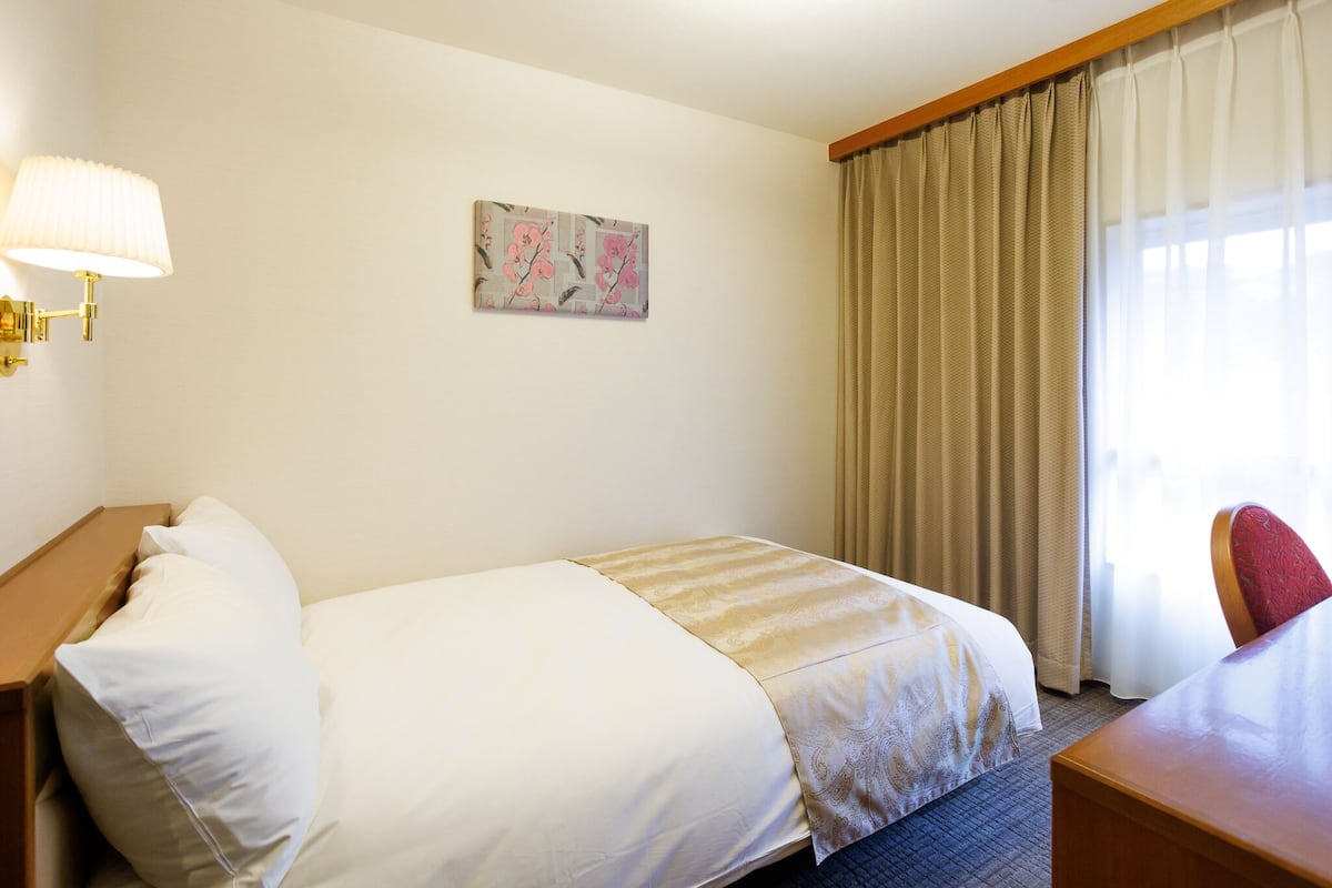 A hotel room with a bed, a desk, a lamp, a painting, and a window with curtains.