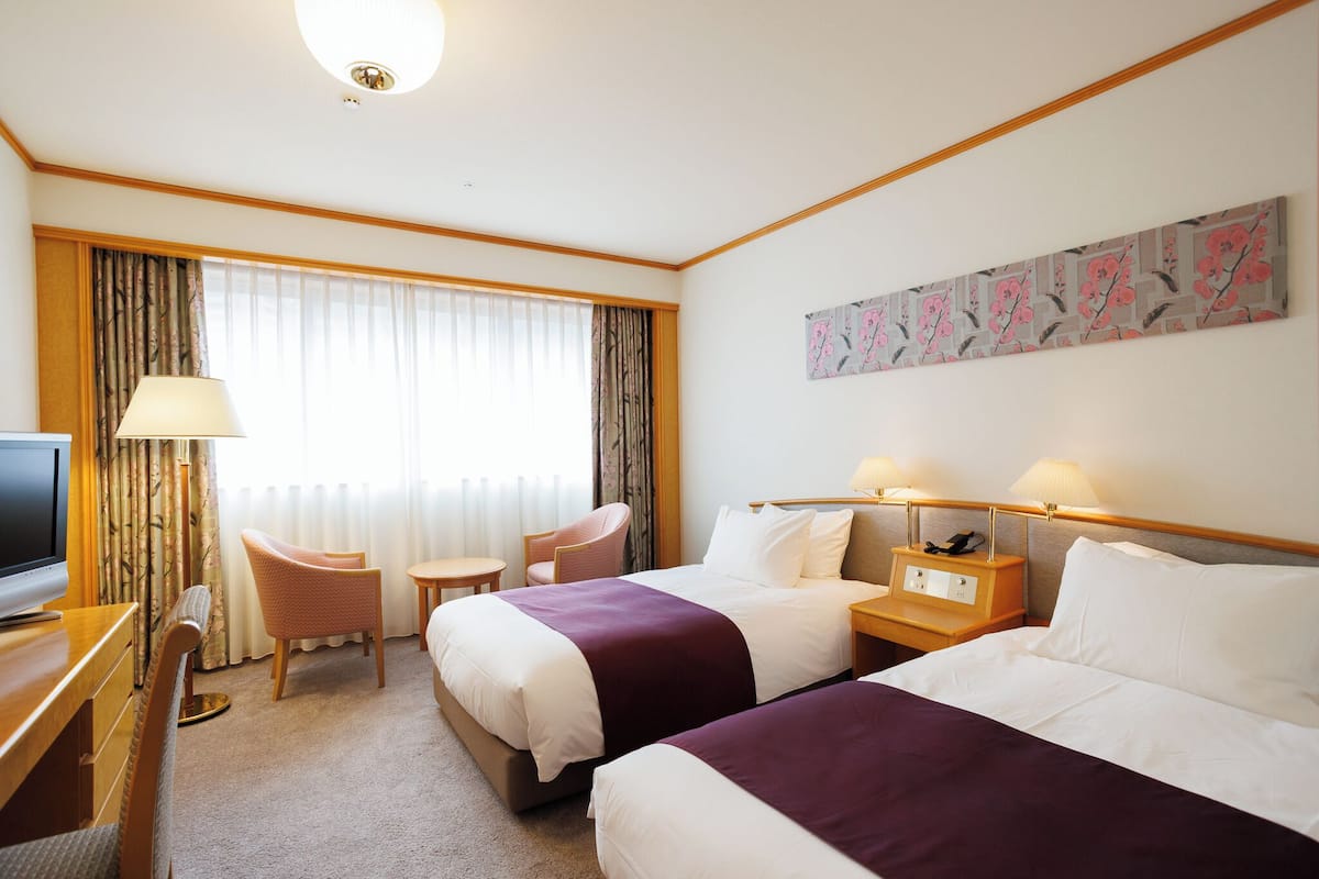 A hotel room with two beds, a desk with a TV, a chair, a small table, and a window with curtains.
