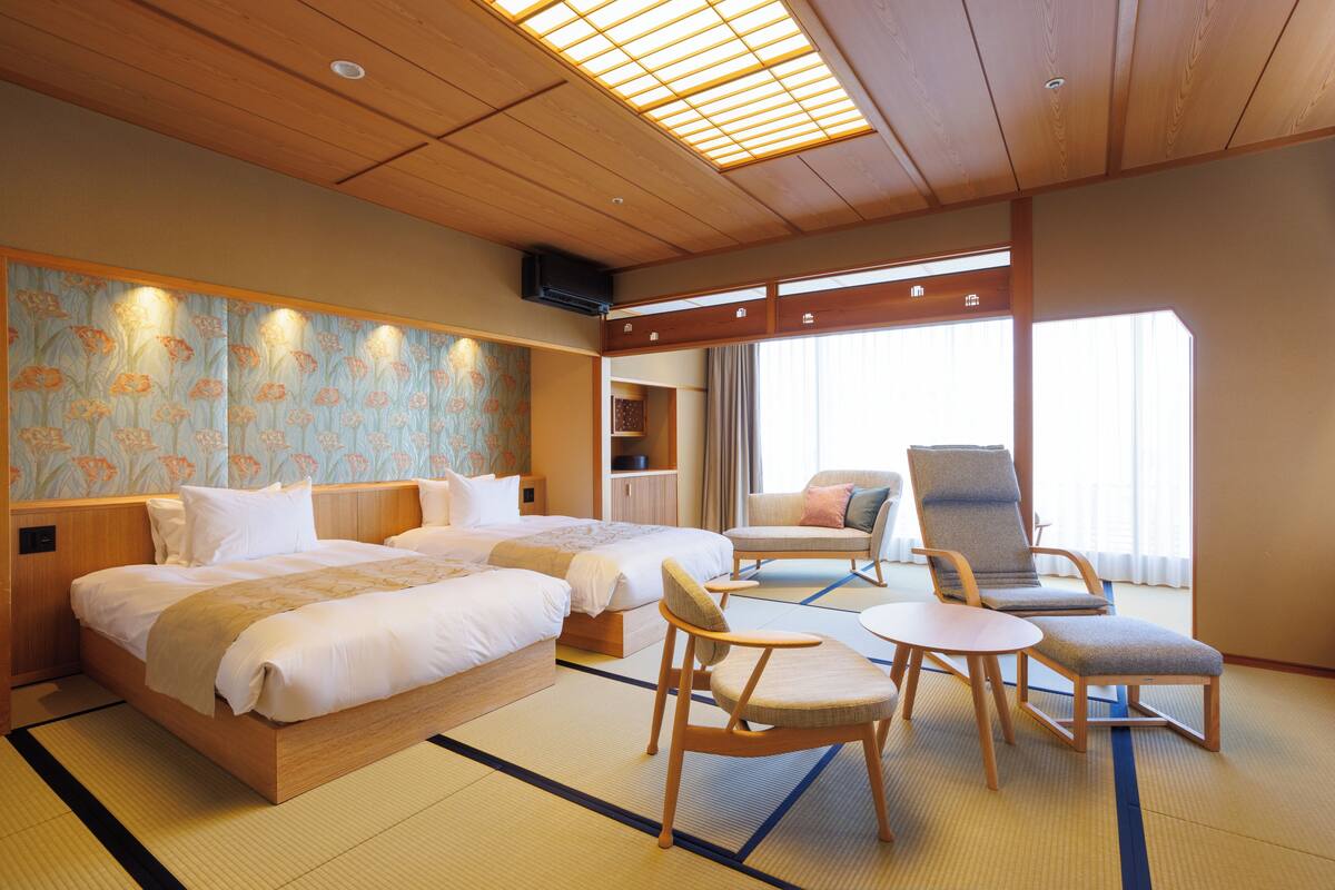 A hotel room with two beds, a sitting area with a chair and table, and a large window with curtains.