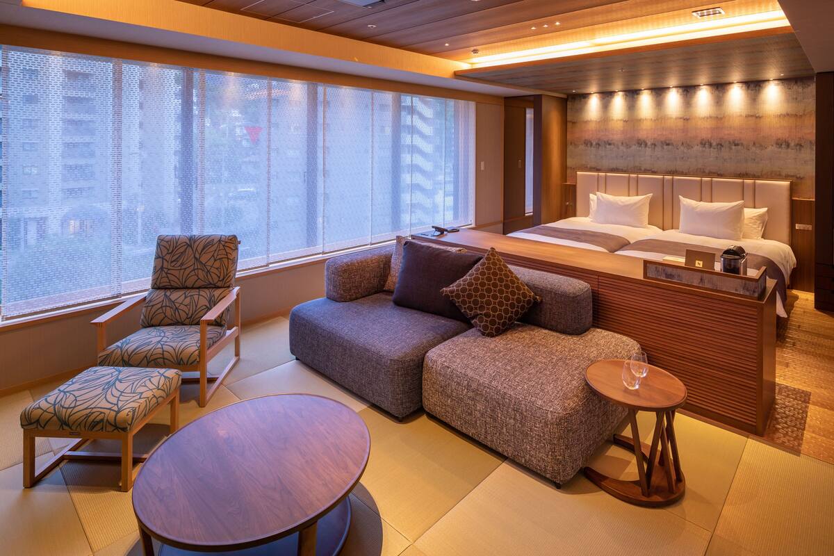 A modern hotel room with a sofa, chairs, a small table, and a bed with a headboard.