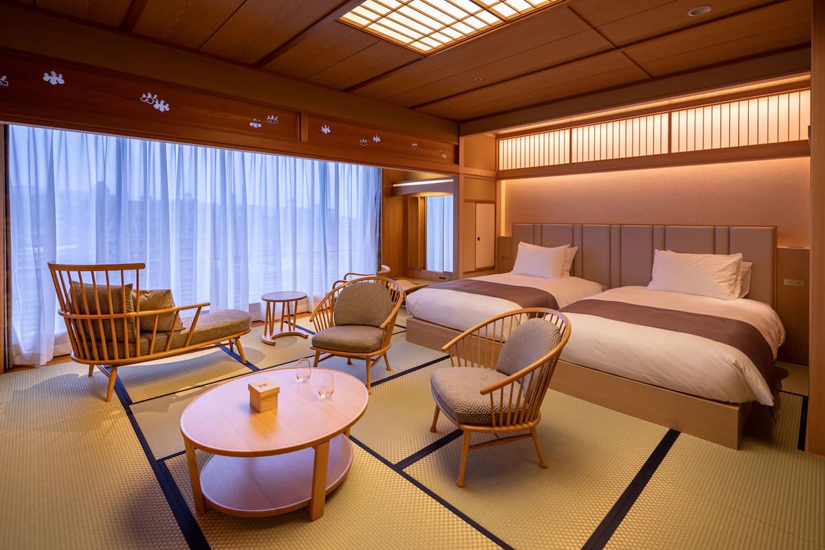 A hotel room with a large bed, two chairs, a small table, and a large window with curtains.