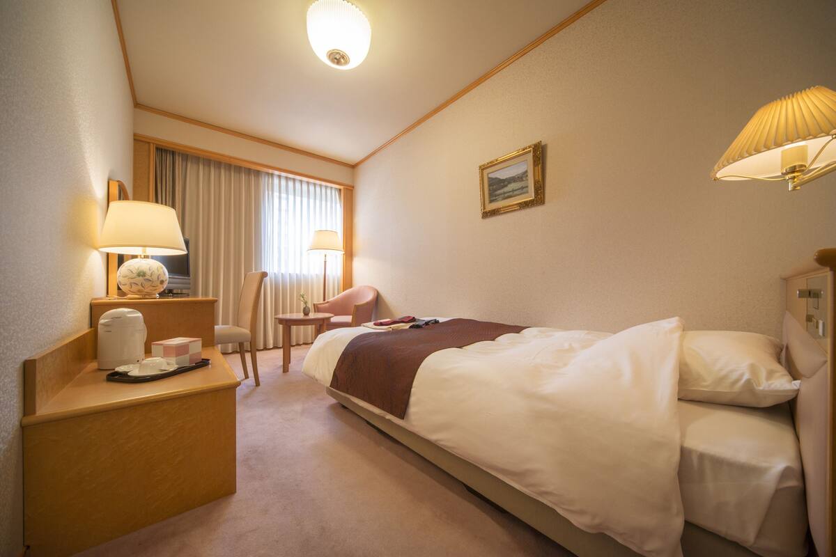 A hotel room with a bed, bedside table, lamp, chair, and a small table with a vase.