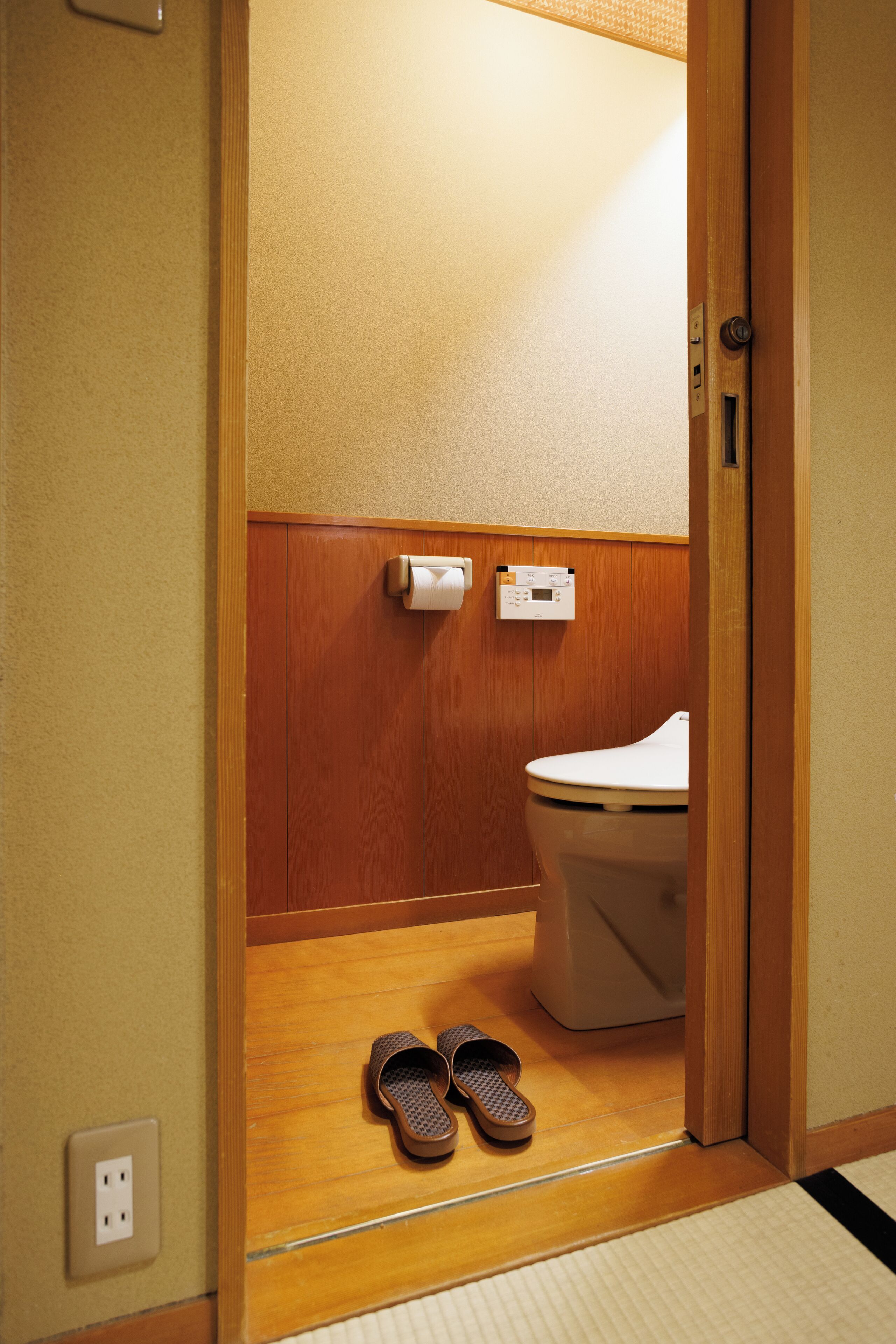 A small, wooden-framed bathroom with a toilet, a paper towel dispenser, and a pair of slippers on the floor.