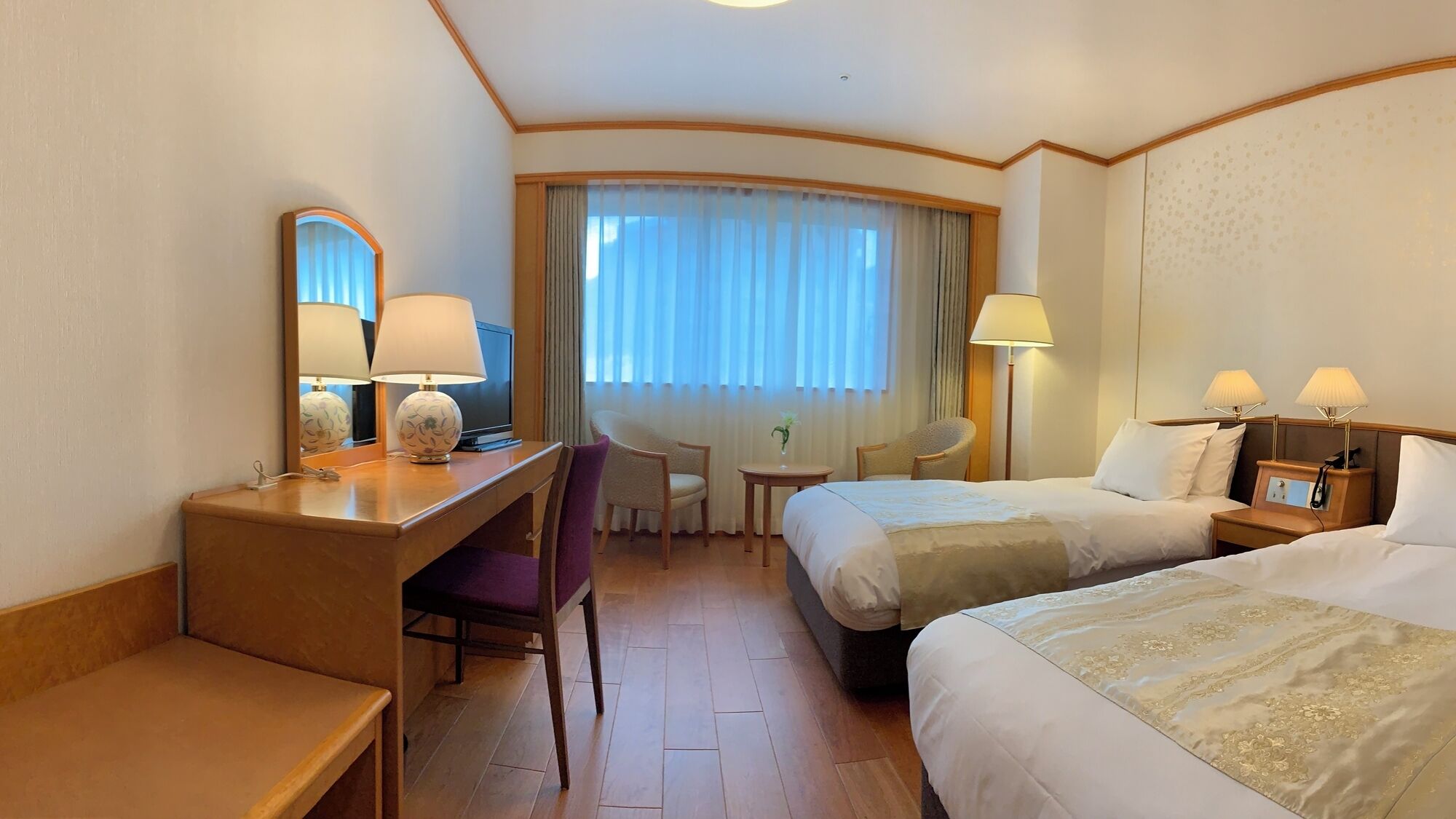 Ein Hotelzimmer mit Schreibtisch, zwei Betten, einem Sessel, einem kleinen Tisch und einem Fenster mit Vorhängen.