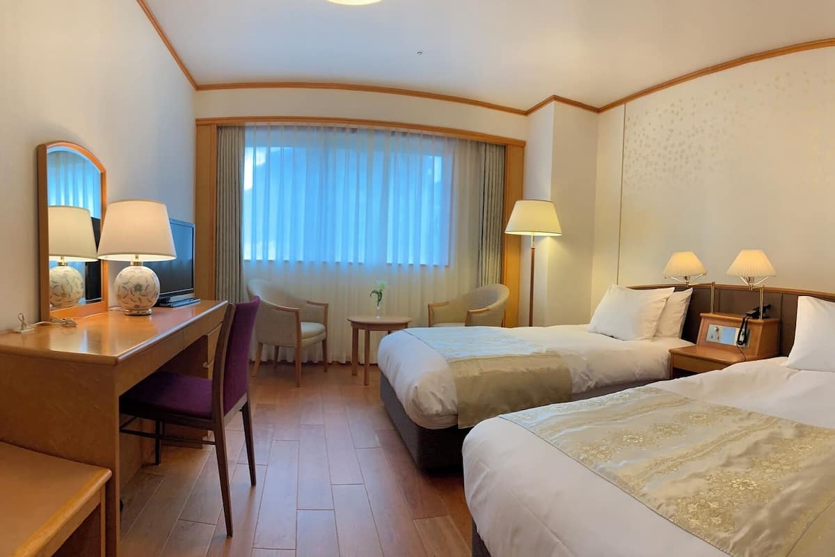 A hotel room with a desk, two beds, a chair, a small table, and a window with curtains.