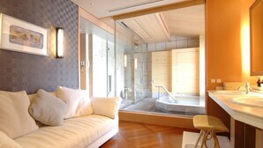 A modern hotel room with a sofa, a small stool, a large mirror, and a bathroom with a bathtub and sink. - Hotel Wakamizu (Takarazuka)