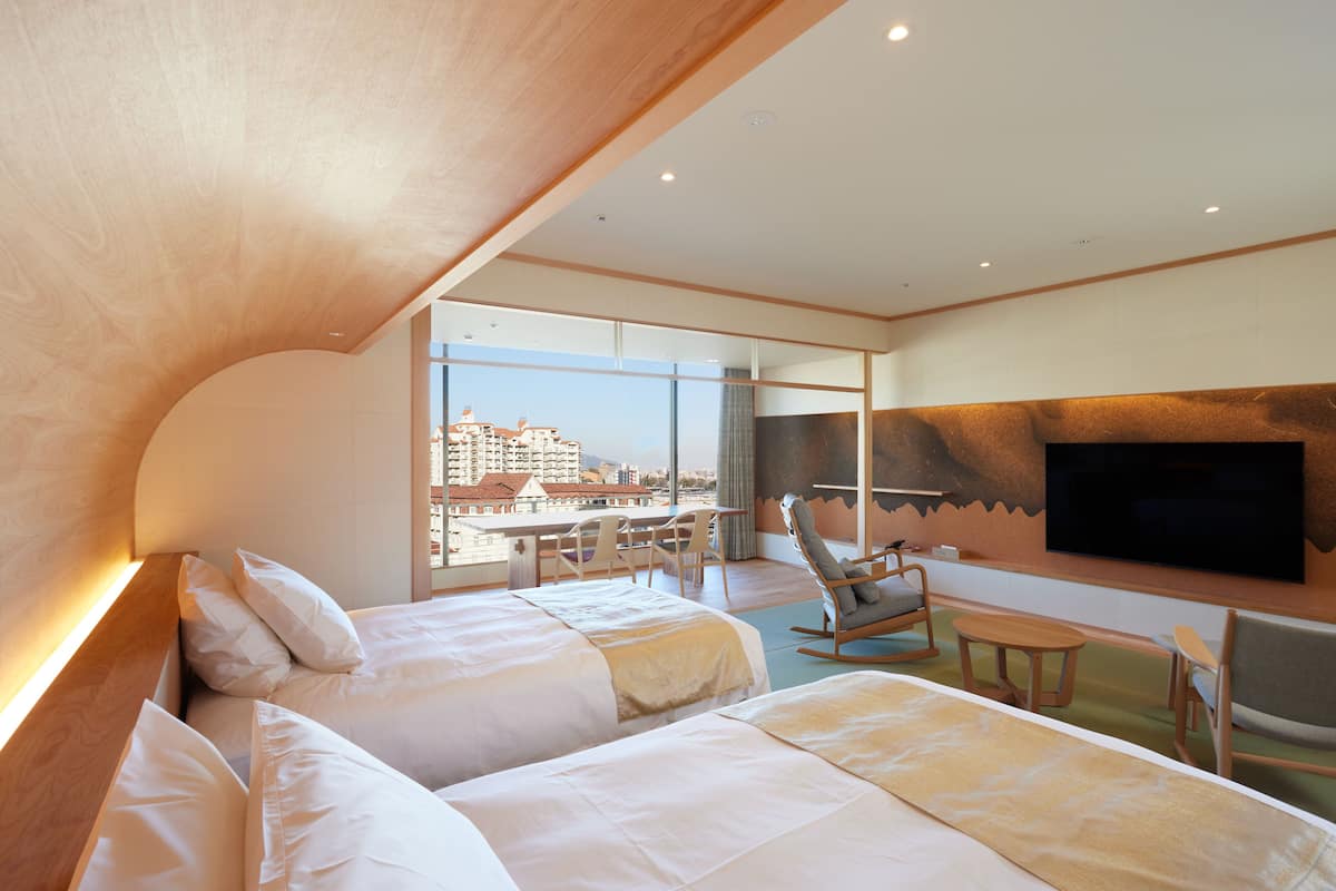 A hotel room with two beds, a TV, a seating area with a table and chairs, and a large window offering a city view.