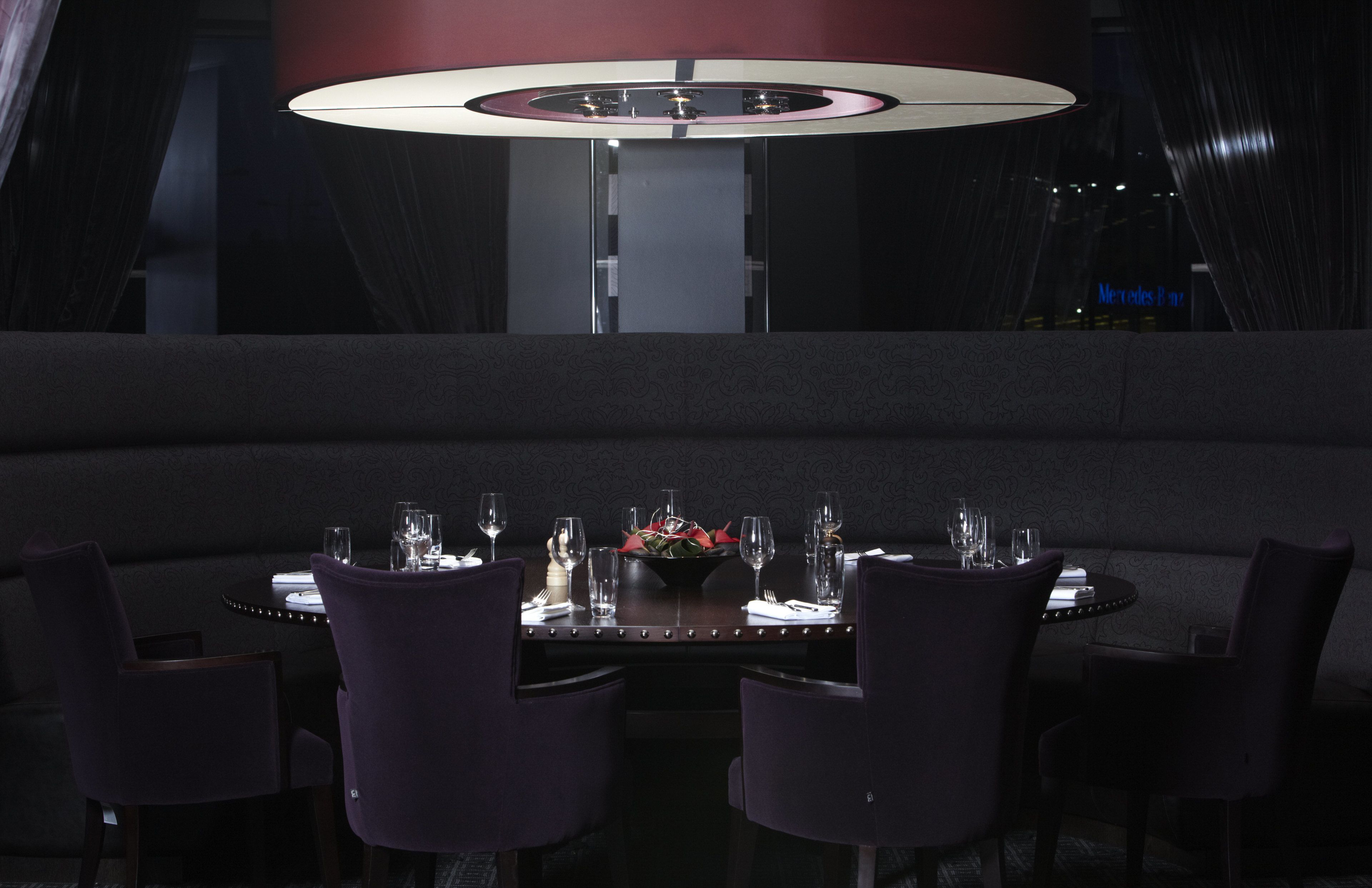 A dimly lit dining area with a round table set for two, surrounded by dark chairs. A central light fixture hangs above the table, and a sign indicating a Mercedes-Benz parking area is visible in the background.