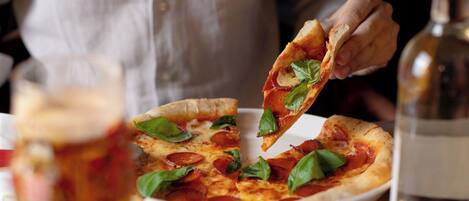 A person holding a slice of pizza with basil leaves, a glass of a beverage, and a bottle on a table.