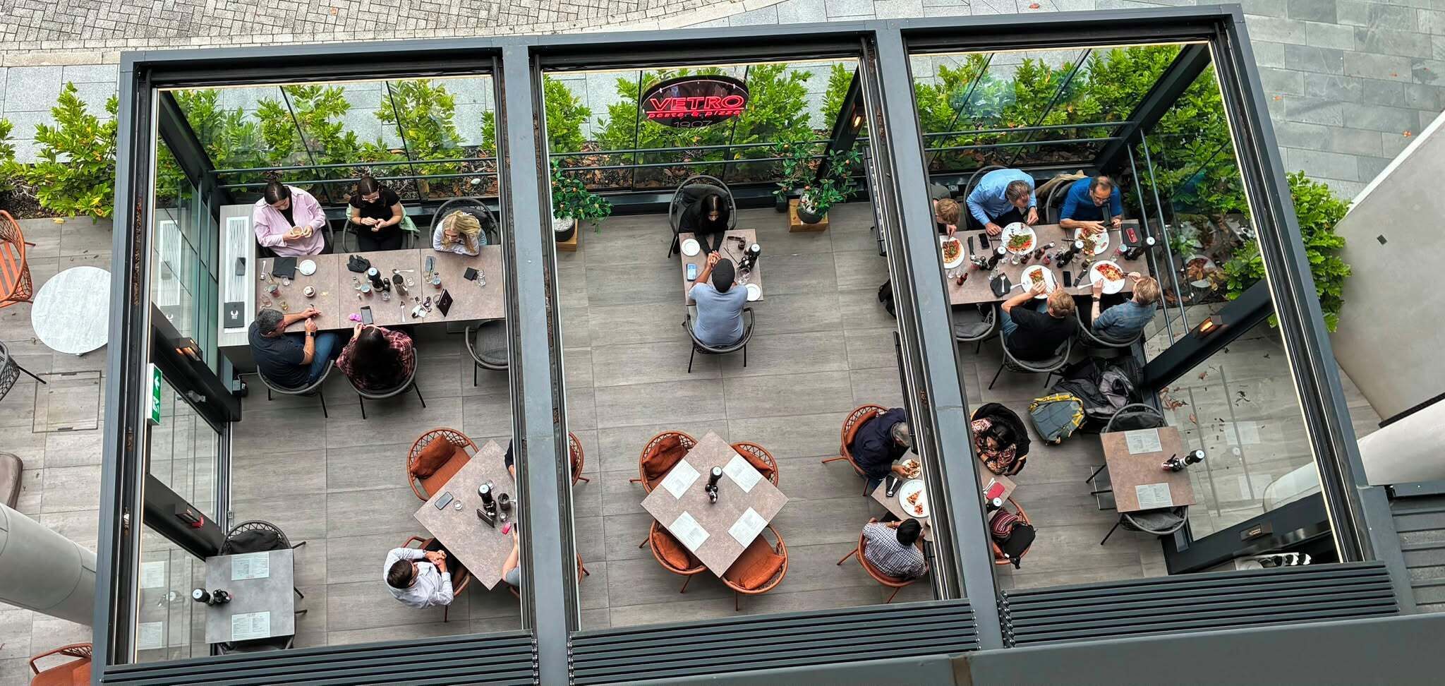 An outdoor dining area with tables and chairs, surrounded by greenery and a glass enclosure.