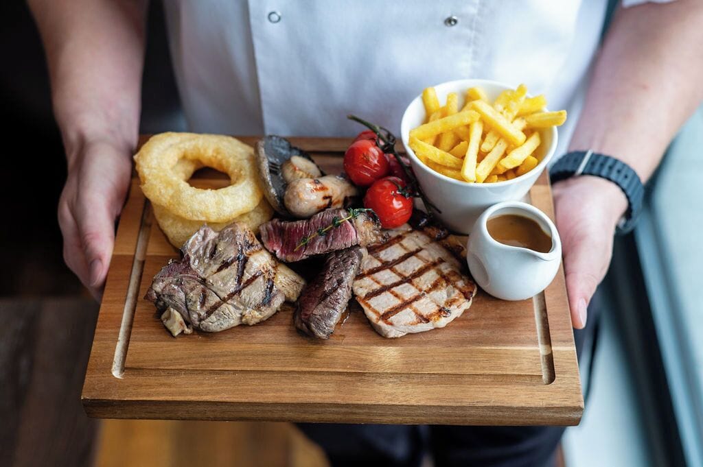 A wooden serving board with grilled meat, onion rings, cherry tomatoes, French fries, and a small jug of sauce.