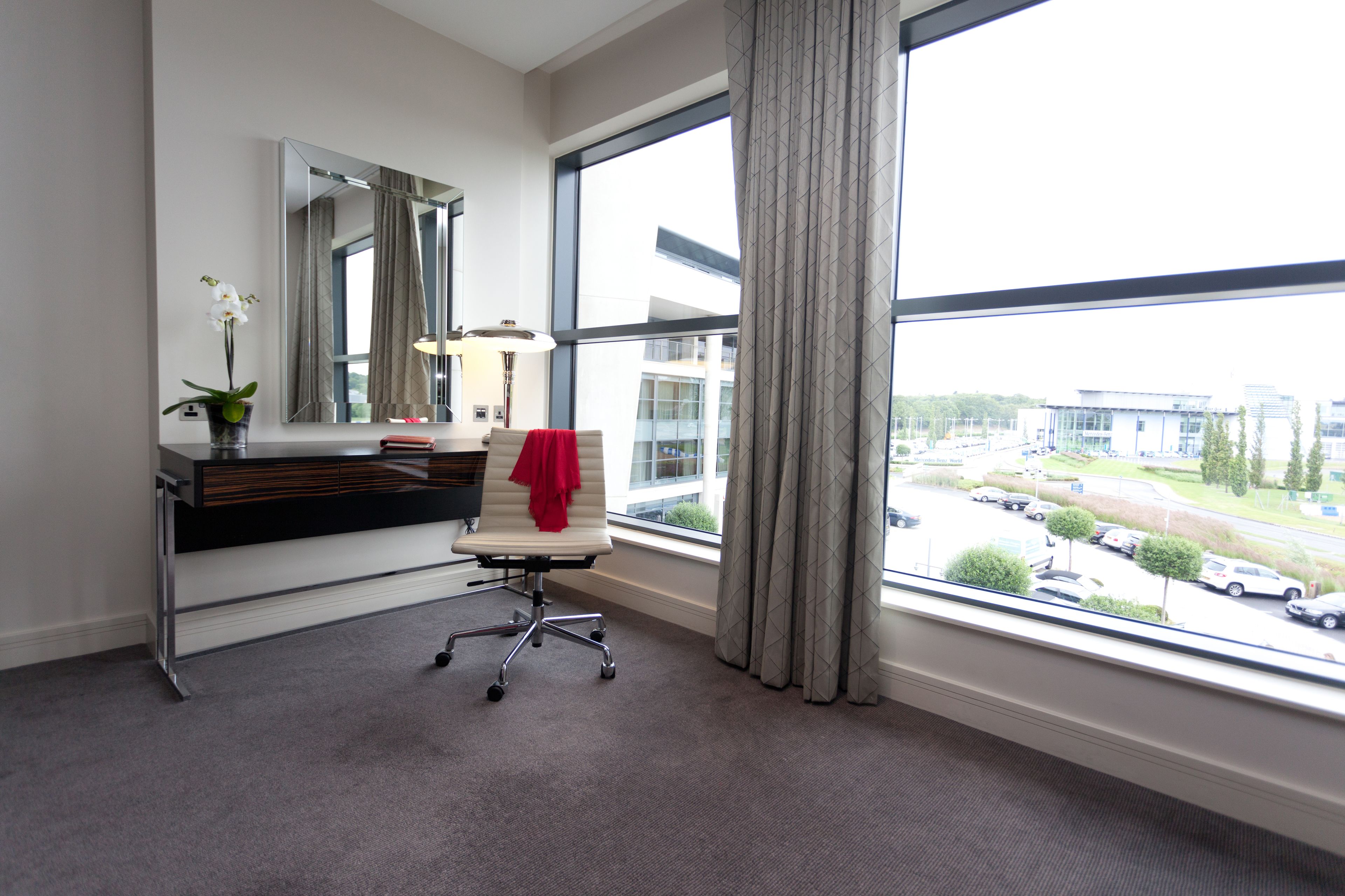 A hotel room with a large window, a desk with a chair, a mirror, and a vase with a plant.